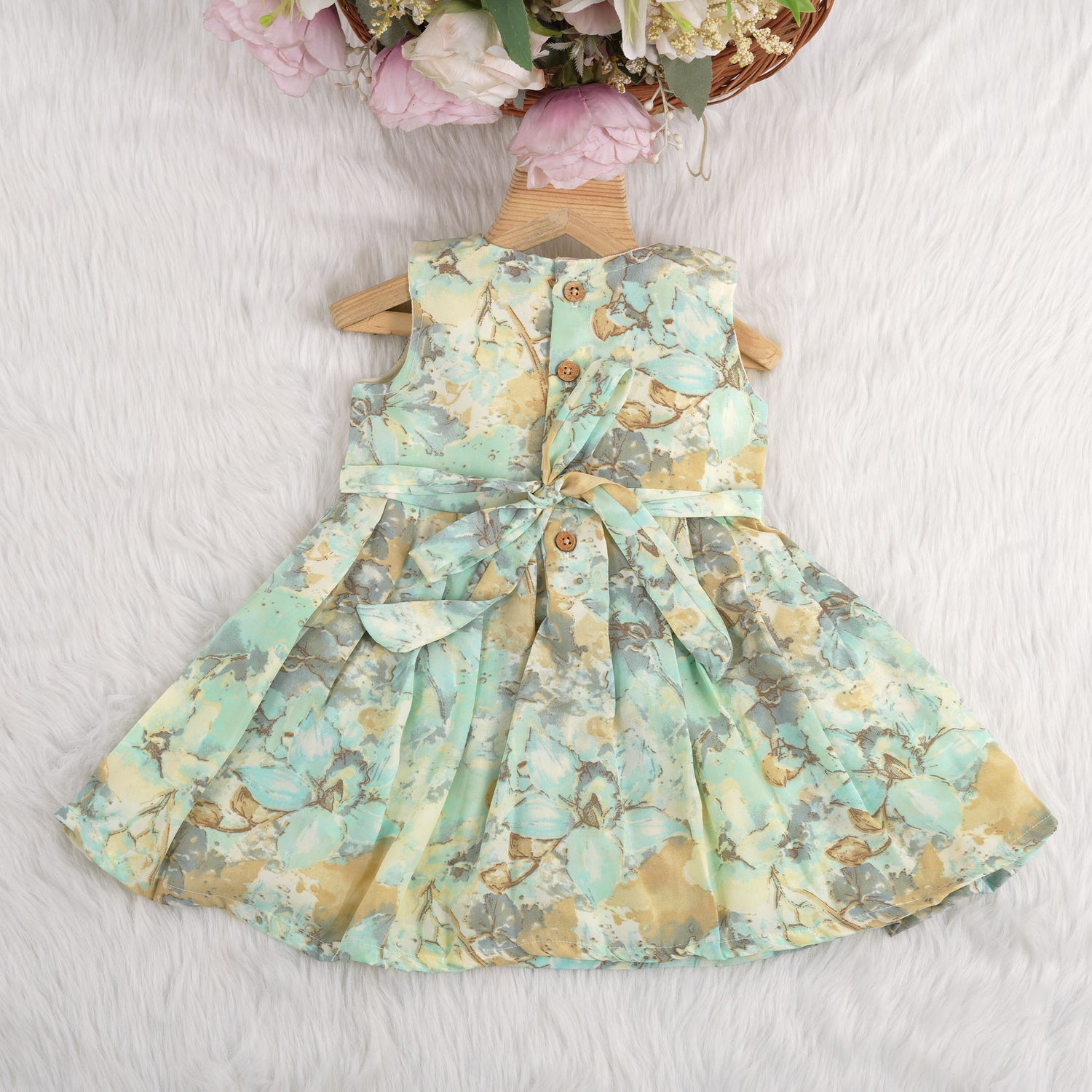 Mint green floral sleeveless chiffon cotton frock for baby girls, pleated and lightweight, displayed on a wooden hanger over a white fur backdrop with pink and white flowers.