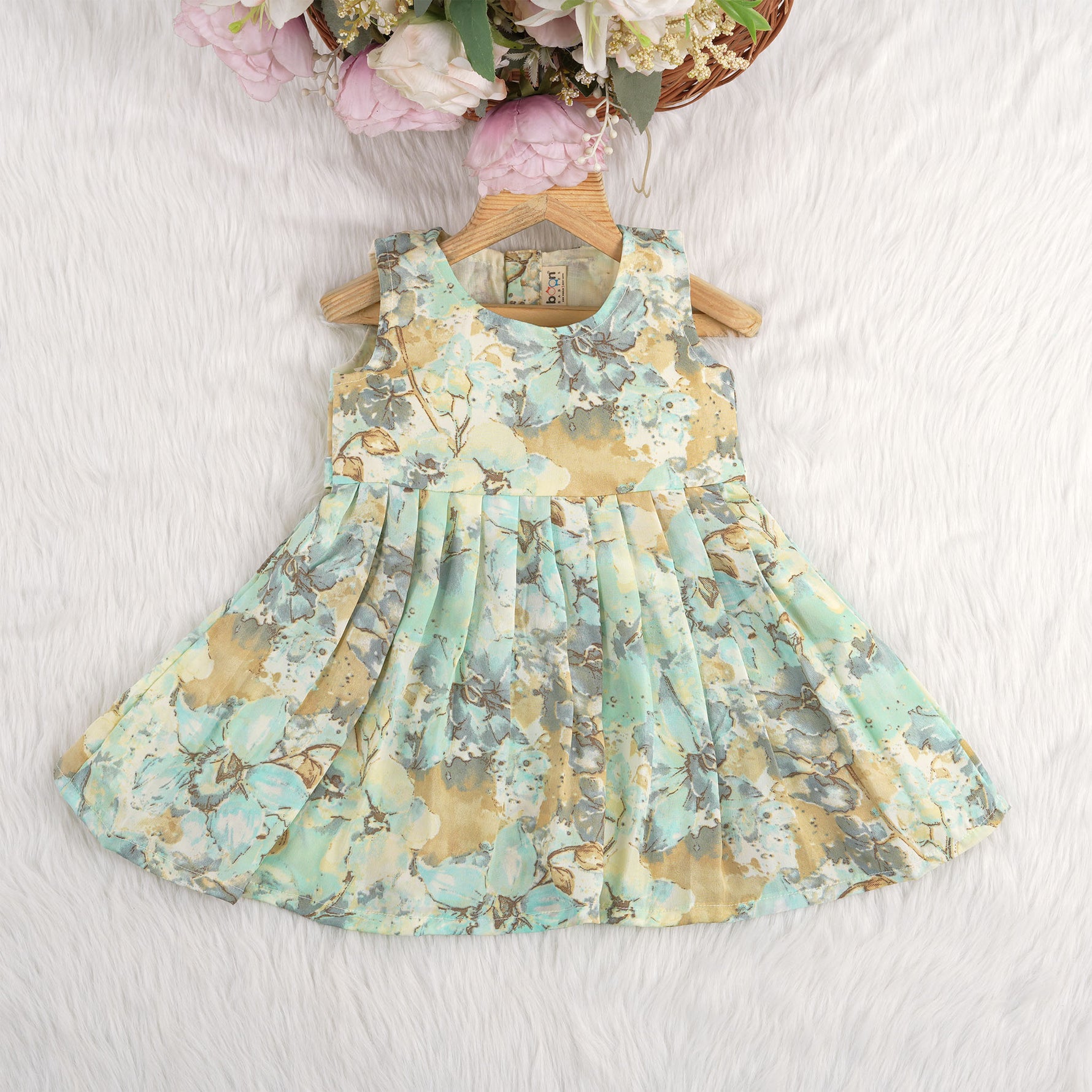 Mint green floral sleeveless chiffon cotton frock for baby girls, pleated and lightweight, displayed on a wooden hanger over a white fur backdrop with pink and white flowers.