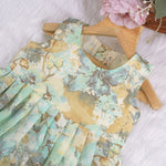 Mint green floral sleeveless chiffon cotton frock for baby girls, pleated and lightweight, displayed on a wooden hanger over a white fur backdrop with pink and white flowers.