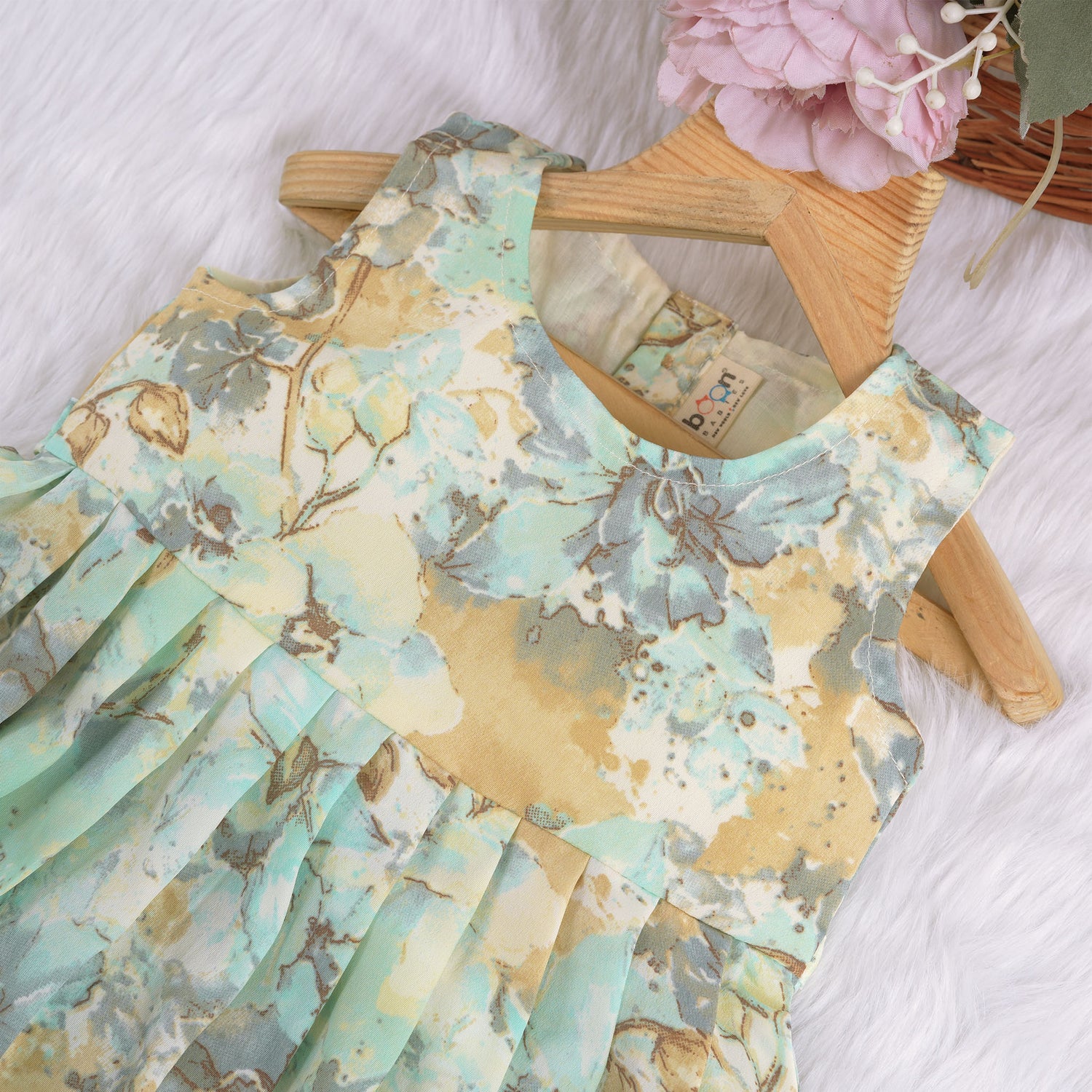 Mint green floral sleeveless chiffon cotton frock for baby girls, pleated and lightweight, displayed on a wooden hanger over a white fur backdrop with pink and white flowers.