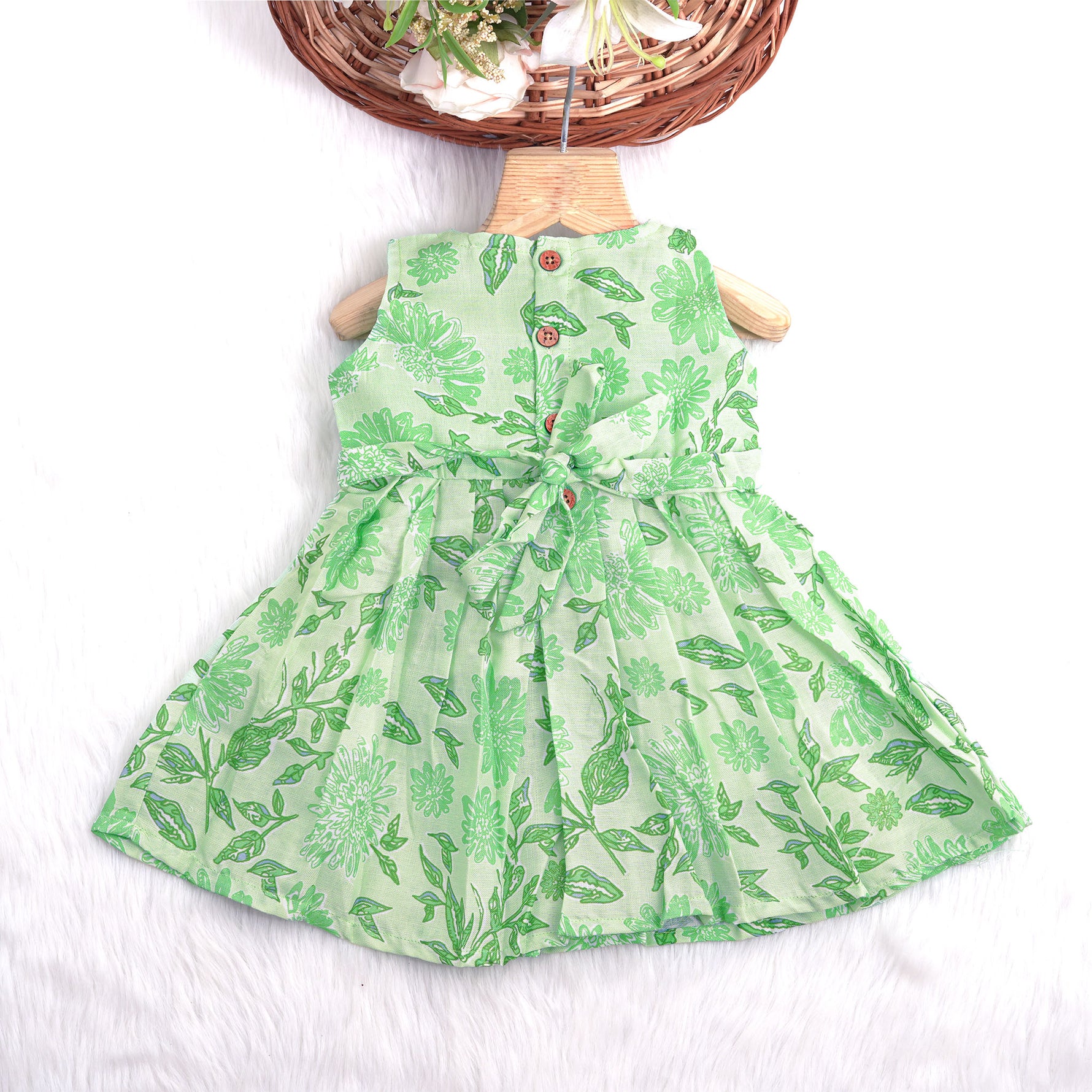 Mint green floral printed sleeveless slub cotton frock for baby girls with pleated flare, displayed flat on white fur background with floral basket hanger.