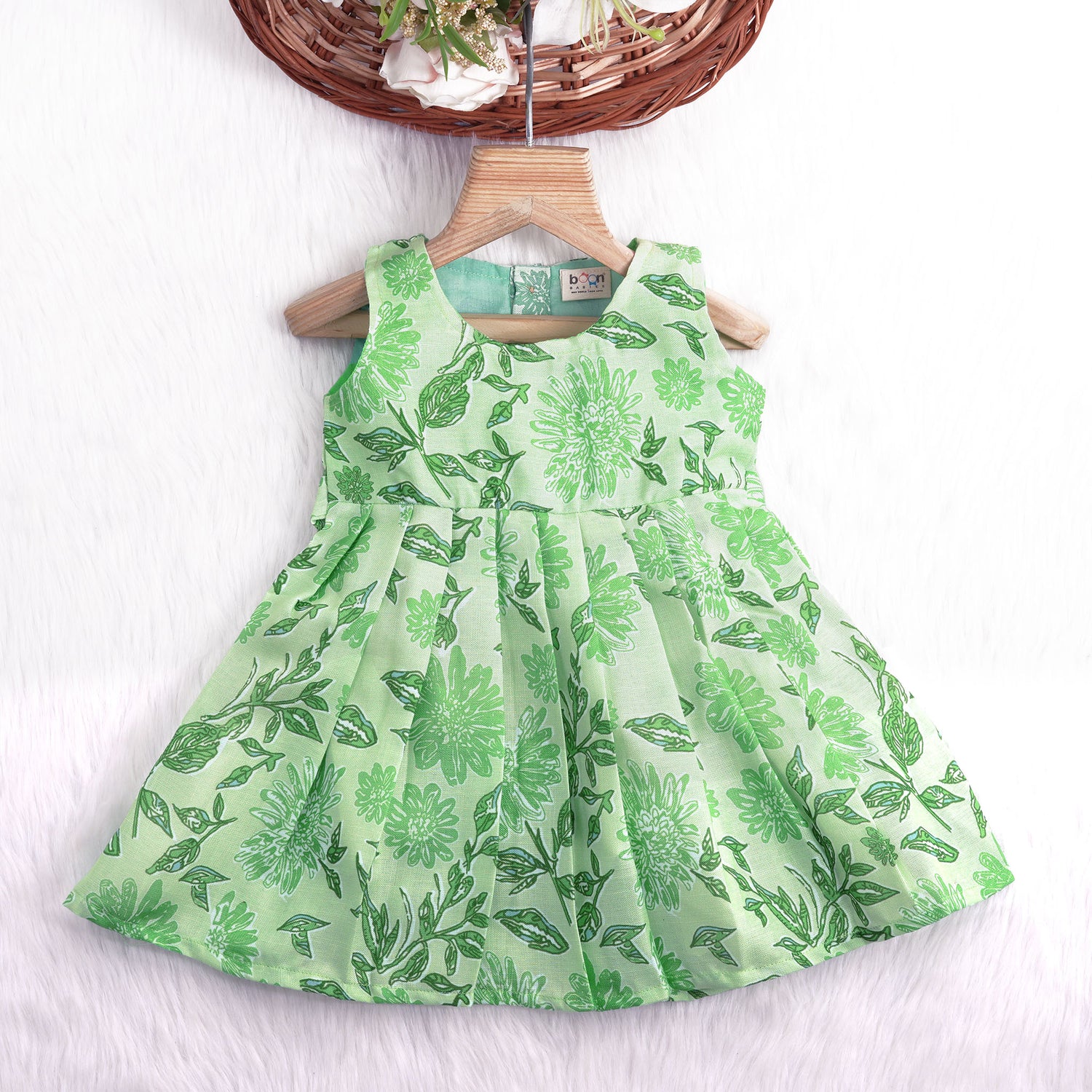 Mint green floral printed sleeveless slub cotton frock for baby girls with pleated flare, displayed flat on white fur background with floral basket hanger.