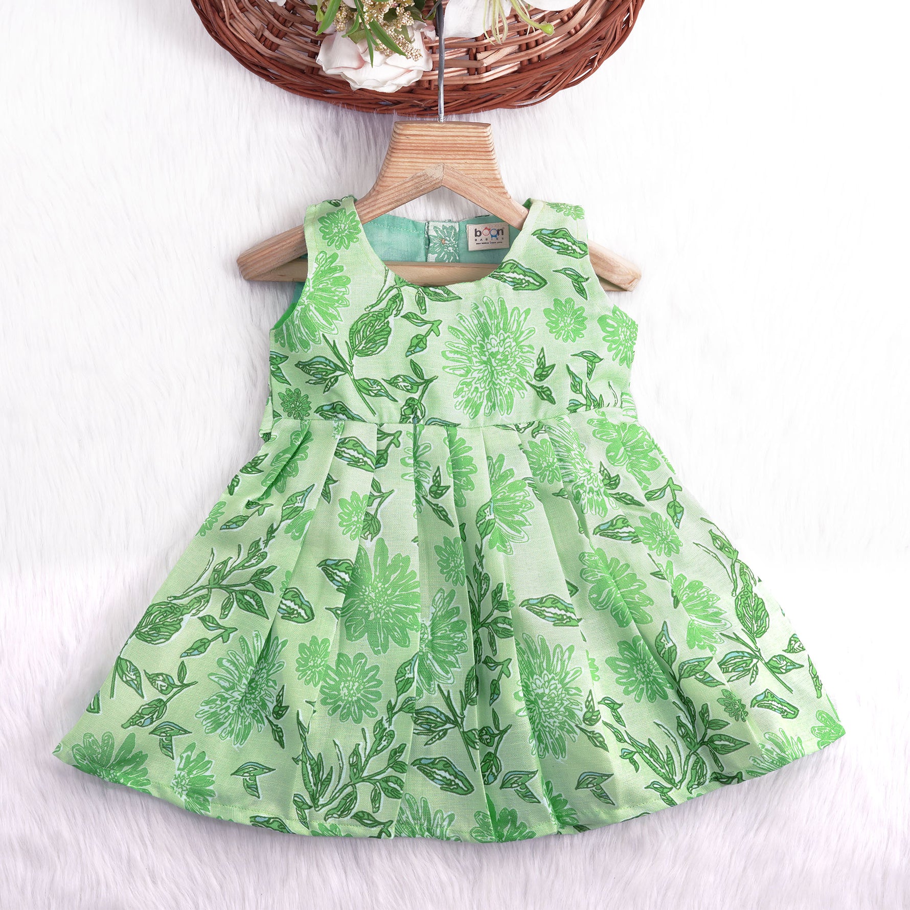 Mint green floral printed sleeveless slub cotton frock for baby girls with pleated flare, displayed flat on white fur background with floral basket hanger.