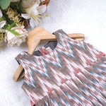 Girls’ sleeveless frock in multicolor Ikat print, made from cotton – displayed flat on white fur background with floral props – Boon Babies casual summer wear for girls aged 12M to 5Y.
