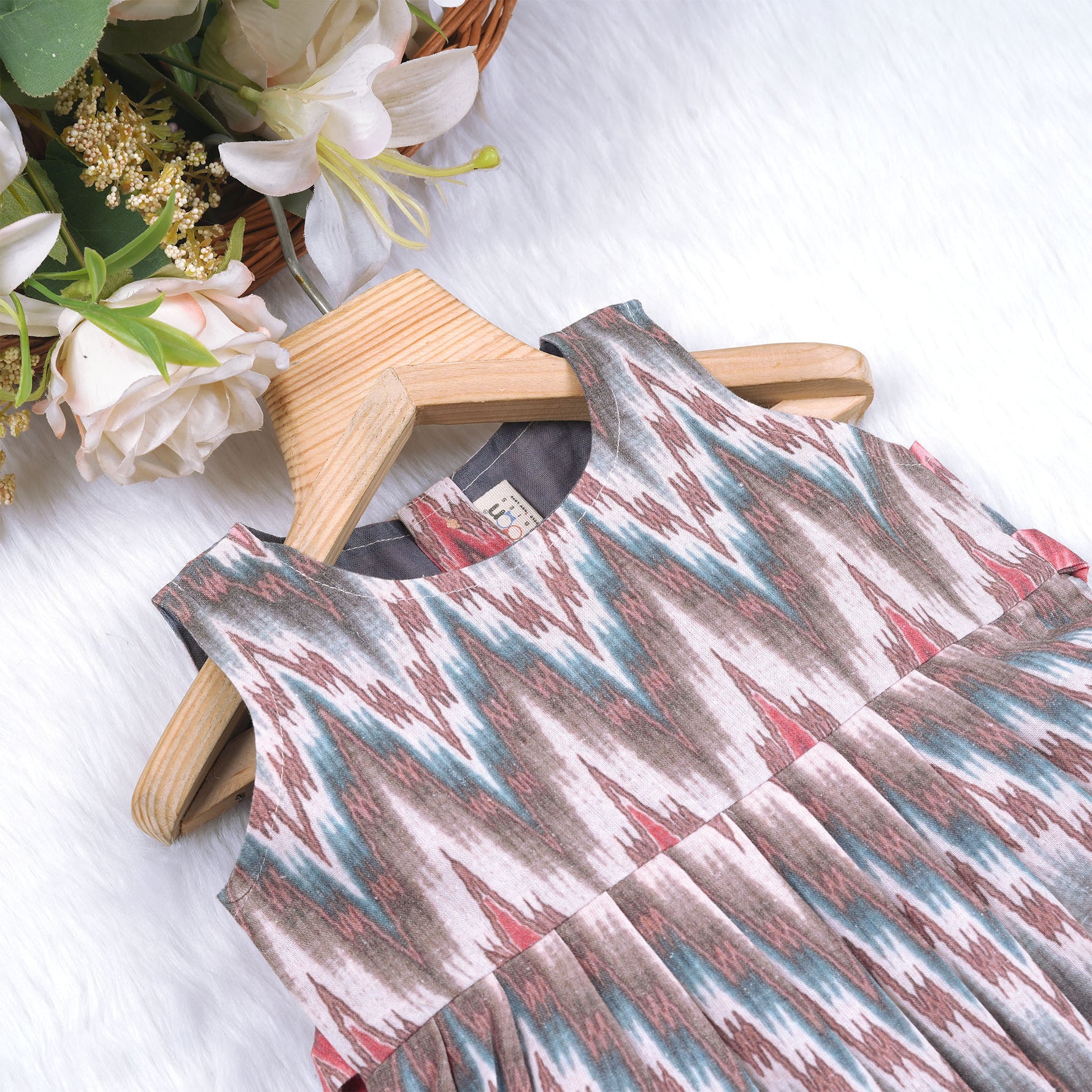Girls’ sleeveless frock in multicolor Ikat print, made from cotton – displayed flat on white fur background with floral props – Boon Babies casual summer wear for girls aged 12M to 5Y.
