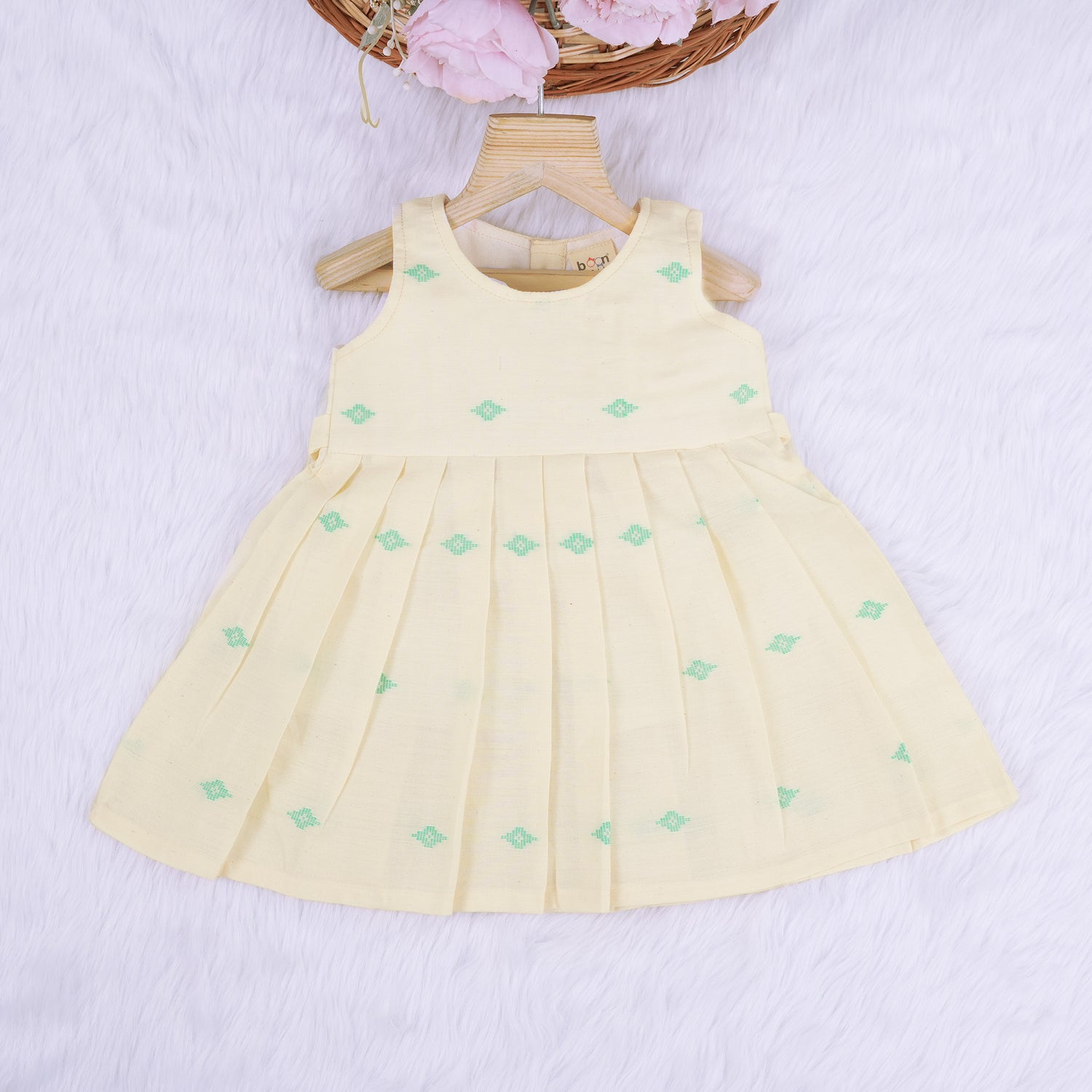 Pastel yellow sleeveless cotton frock with mint green embroidery and pleated skirt for baby girls, displayed on a white fur background with floral basket hanger.