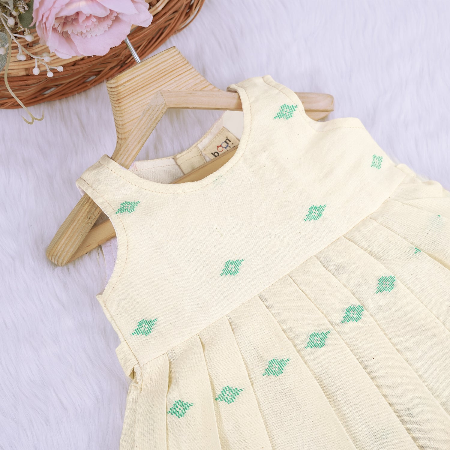 Pastel yellow sleeveless cotton frock with mint green embroidery and pleated skirt for baby girls, displayed on a white fur background with floral basket hanger.