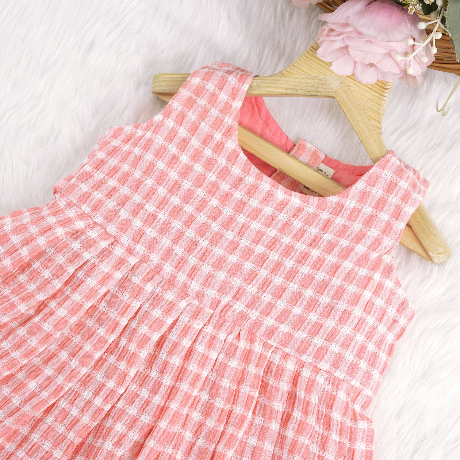 Peach and white checked sleeveless crinkled cotton frock for baby girls, featuring pleated skirt, displayed on a wooden hanger over a white fur background with flowers
