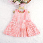 Peach and white checked sleeveless crinkled cotton frock for baby girls, featuring pleated skirt, displayed on a wooden hanger over a white fur background with flowers