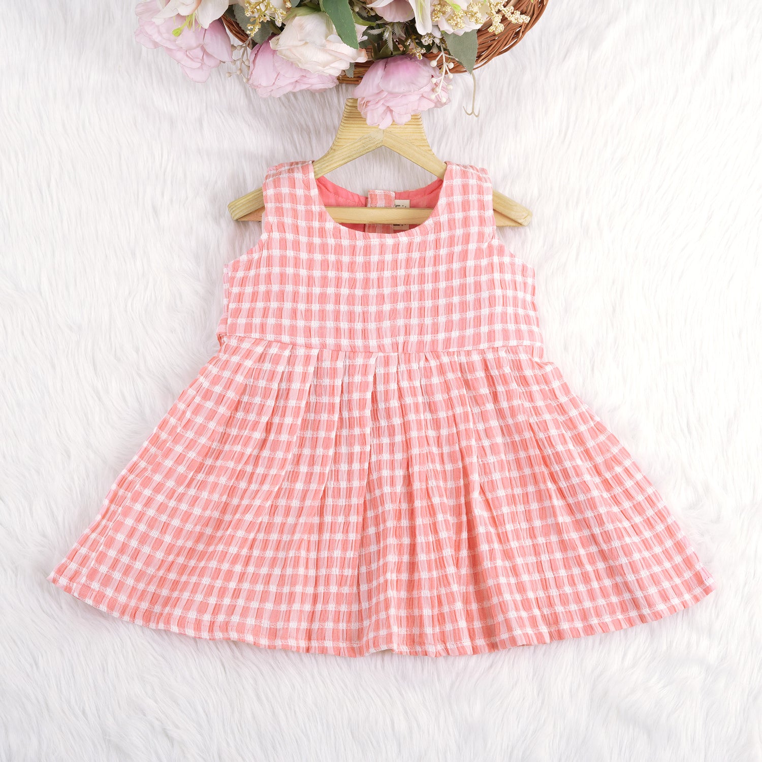 Peach and white checked sleeveless crinkled cotton frock for baby girls, featuring pleated skirt, displayed on a wooden hanger over a white fur background with flowers