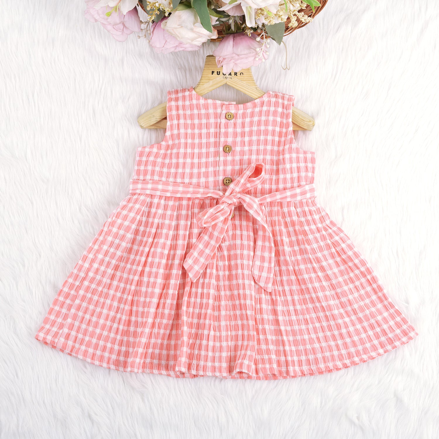 Peach and white checked sleeveless crinkled cotton frock for baby girls, featuring pleated skirt, displayed on a wooden hanger over a white fur background with flowers