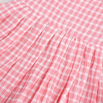 Peach and white checked sleeveless crinkled cotton frock for baby girls, featuring pleated skirt, displayed on a wooden hanger over a white fur background with flowers
