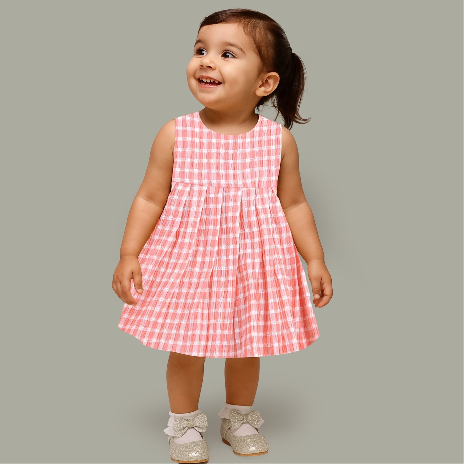 Peach and white checked sleeveless crinkled cotton frock for baby girls, featuring pleated skirt, displayed on a wooden hanger over a white fur background with flowers.