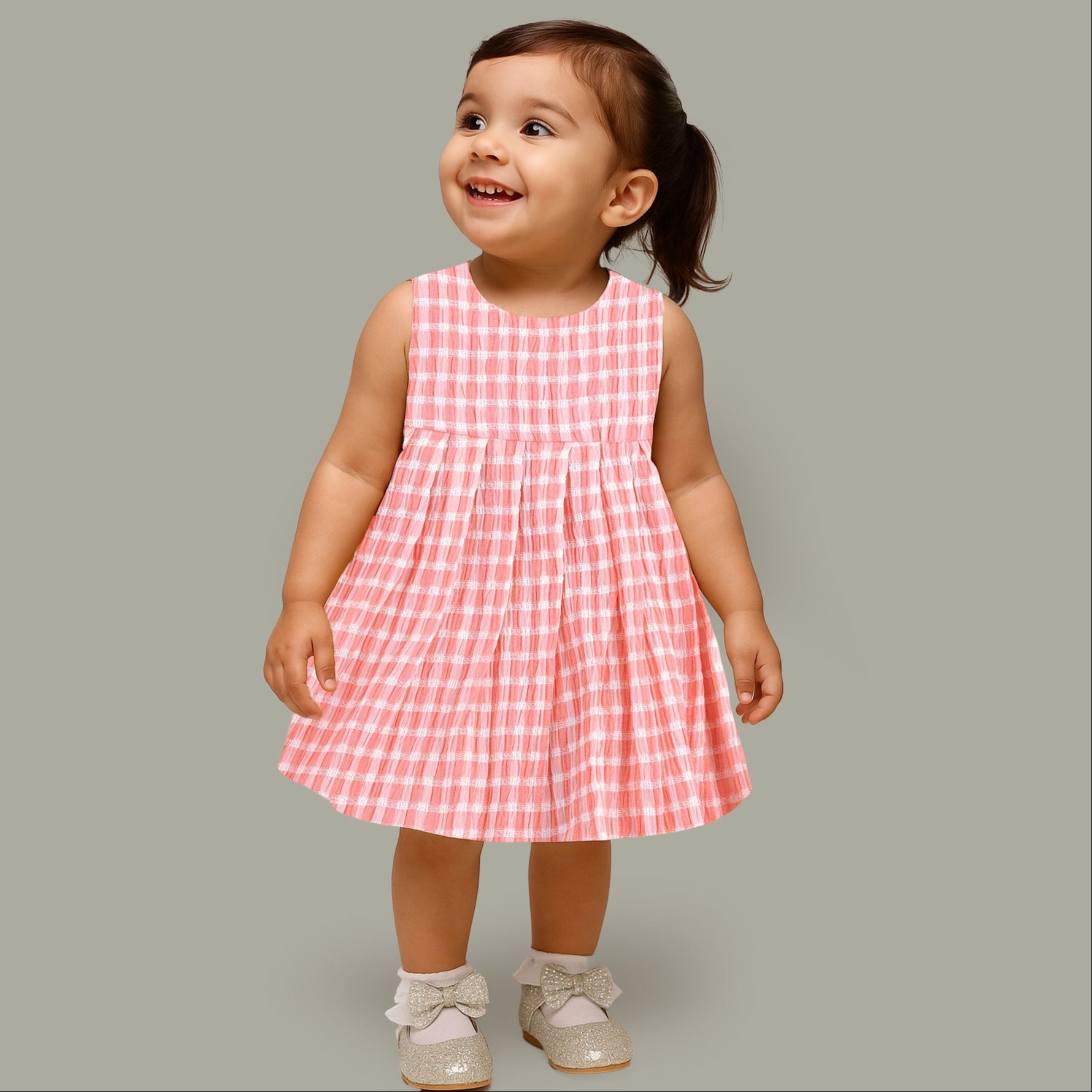 Peach and white checked sleeveless crinkled cotton frock for baby girls, featuring pleated skirt, displayed on a wooden hanger over a white fur background with flowers.