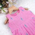 Pink sleeveless cotton frock with soft pleats and blue ikat motifs, displayed on a wooden hanger under a basket of white flowers – from Boon Babies girlswear collection.
