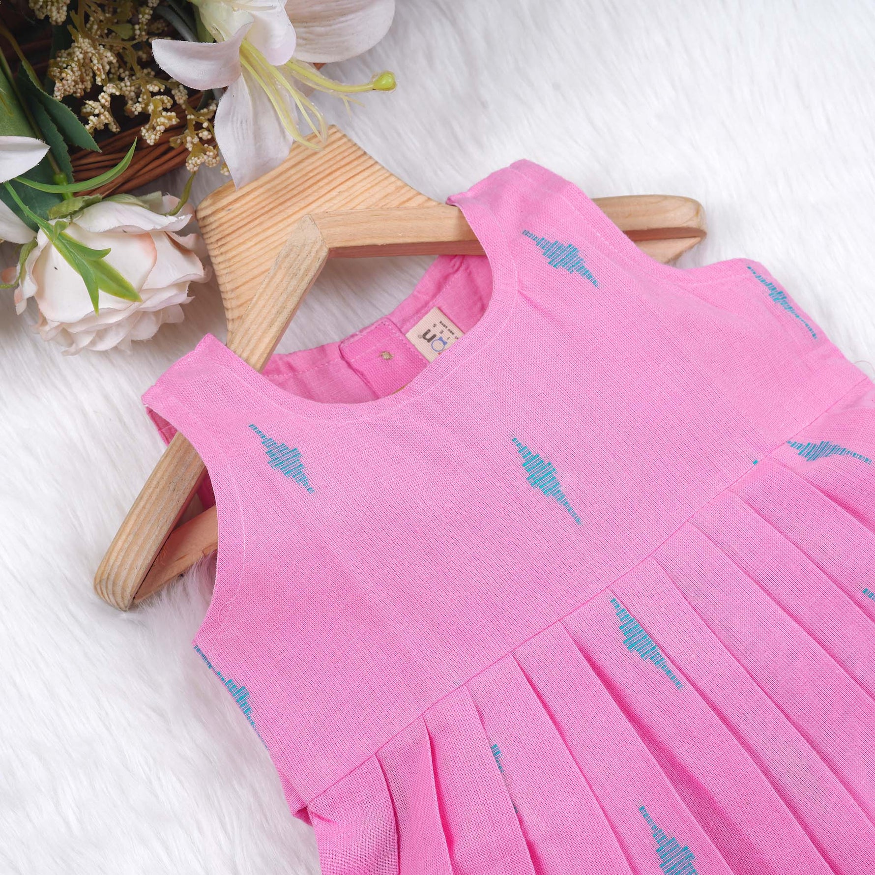 Pink sleeveless cotton frock with soft pleats and blue ikat motifs, displayed on a wooden hanger under a basket of white flowers – from Boon Babies girlswear collection.

