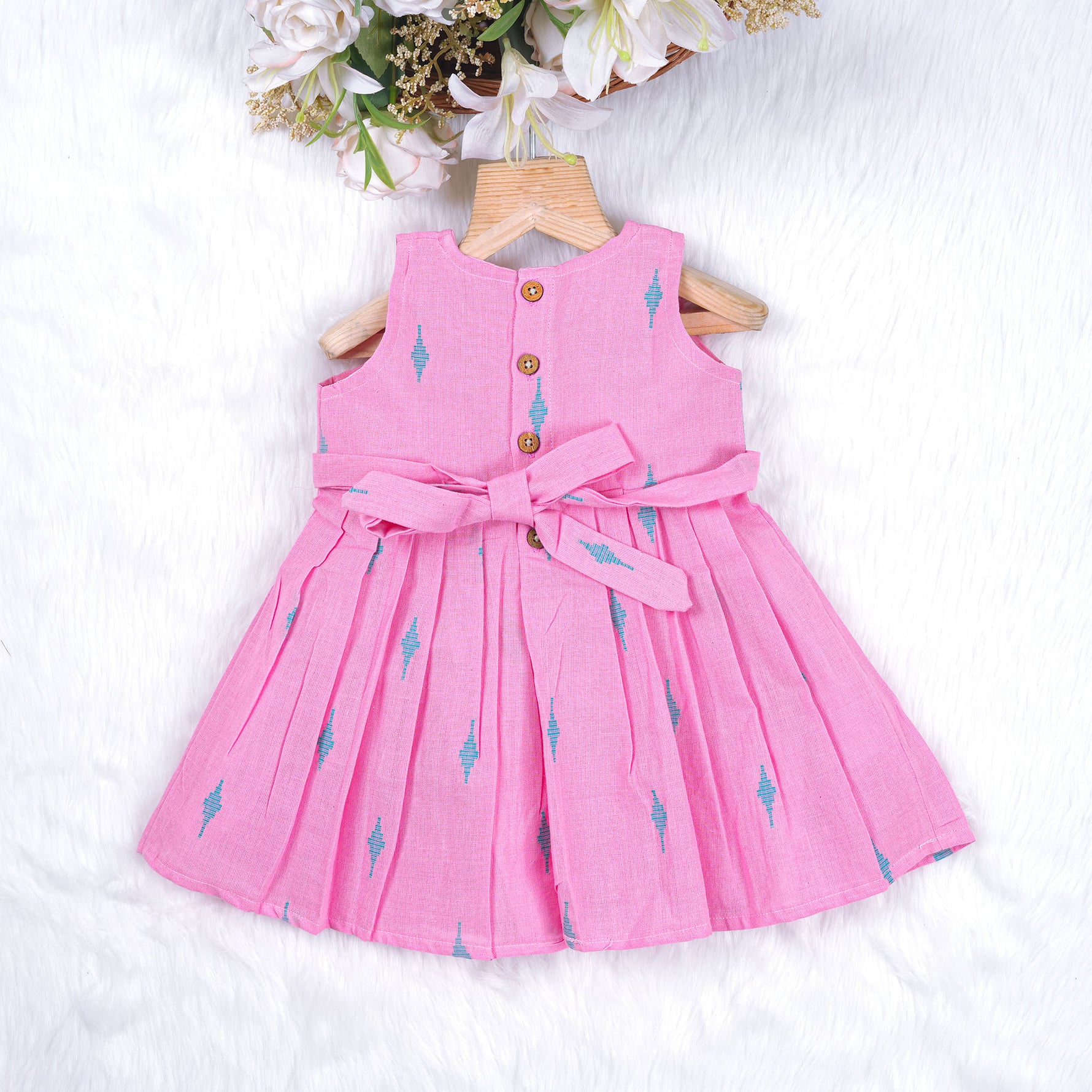 Pink sleeveless cotton frock with soft pleats and blue ikat motifs, displayed on a wooden hanger under a basket of white flowers – from Boon Babies girlswear collection.
