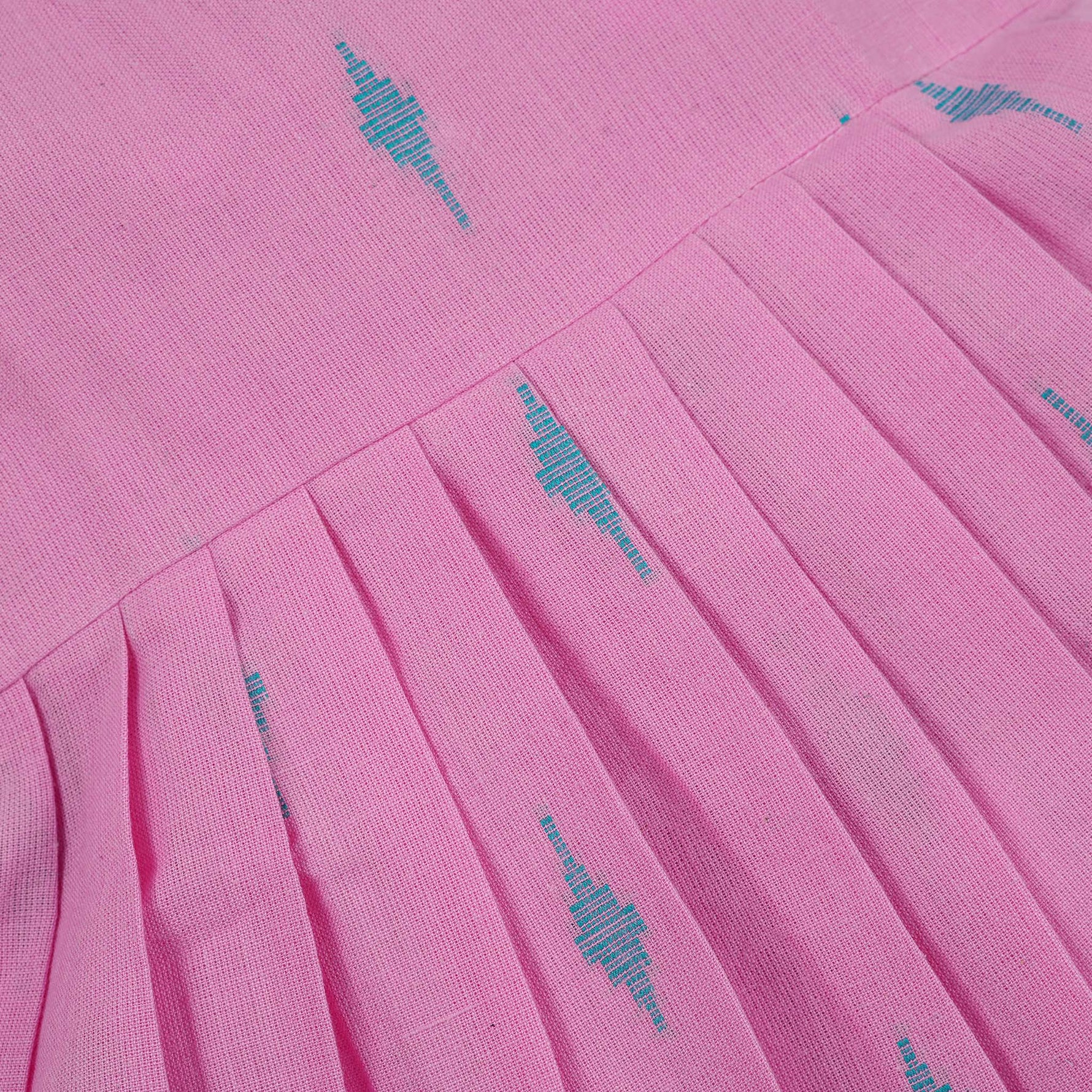 Pink sleeveless cotton frock with soft pleats and blue ikat motifs, displayed on a wooden hanger under a basket of white flowers – from Boon Babies girlswear collection.
