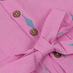 Pink sleeveless cotton frock with soft pleats and blue ikat motifs, displayed on a wooden hanger under a basket of white flowers – from Boon Babies girlswear collection.
