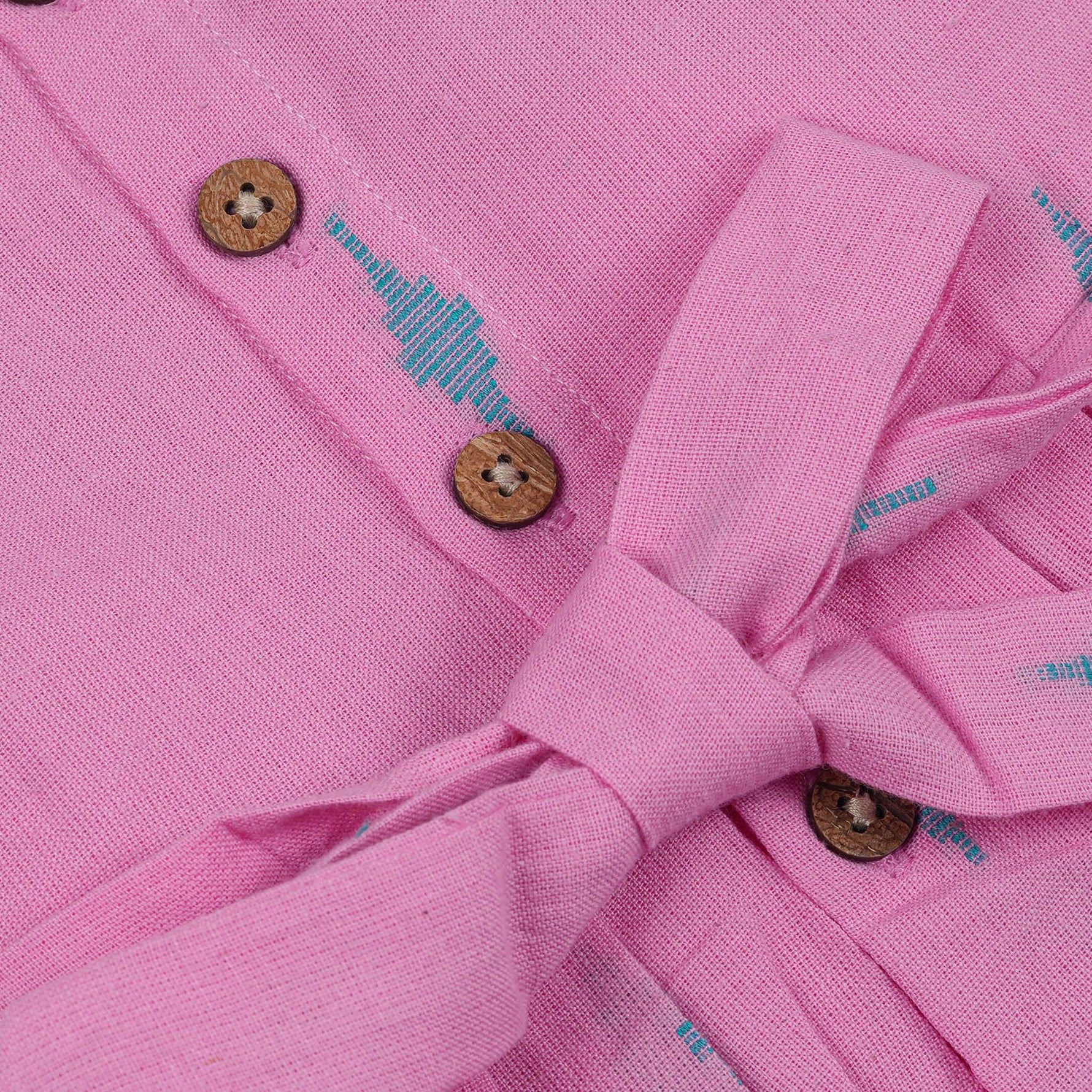 Pink sleeveless cotton frock with soft pleats and blue ikat motifs, displayed on a wooden hanger under a basket of white flowers – from Boon Babies girlswear collection.
