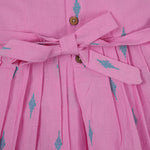 Pink sleeveless cotton frock with soft pleats and blue ikat motifs, displayed on a wooden hanger under a basket of white flowers – from Boon Babies girlswear collection.
