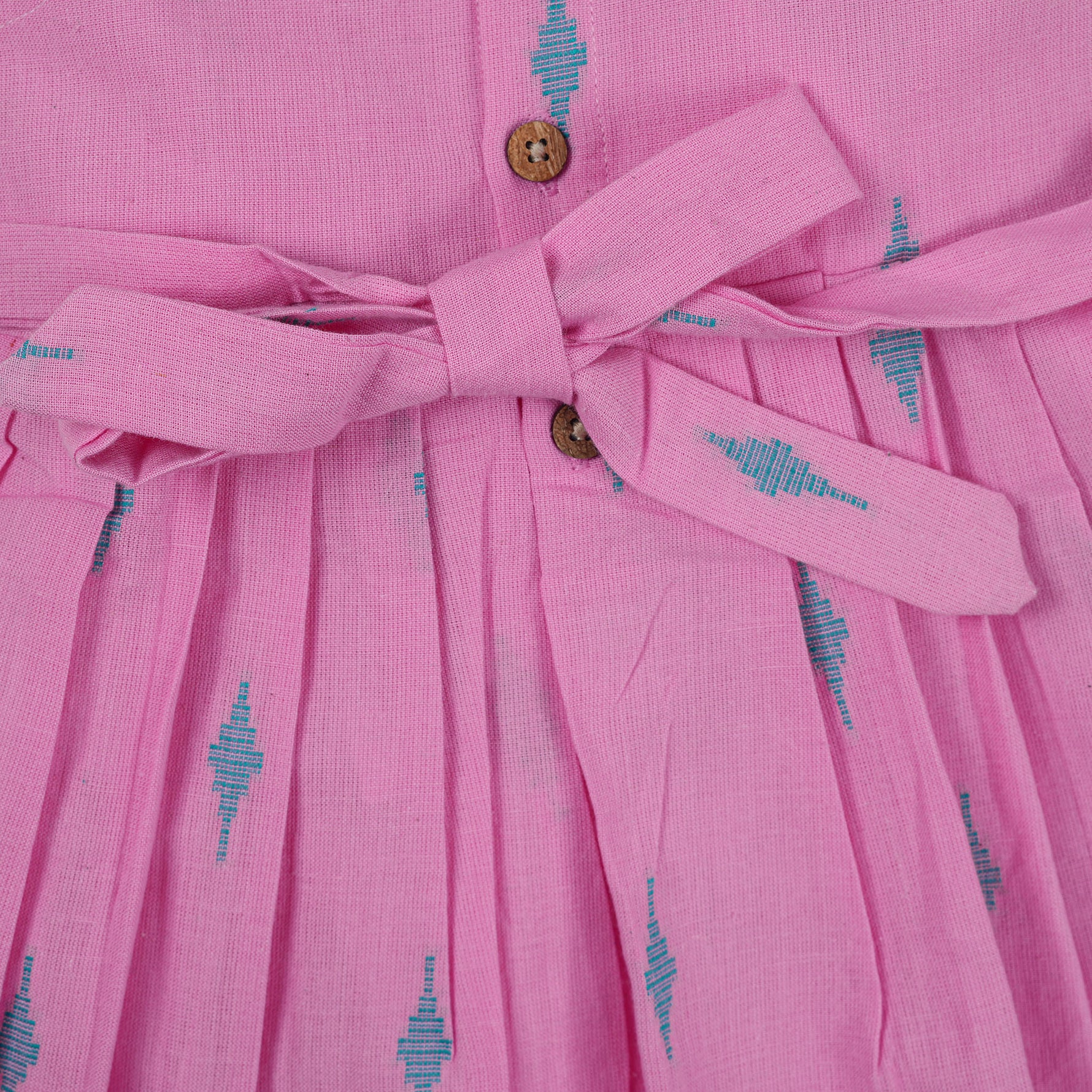 Pink sleeveless cotton frock with soft pleats and blue ikat motifs, displayed on a wooden hanger under a basket of white flowers – from Boon Babies girlswear collection.
