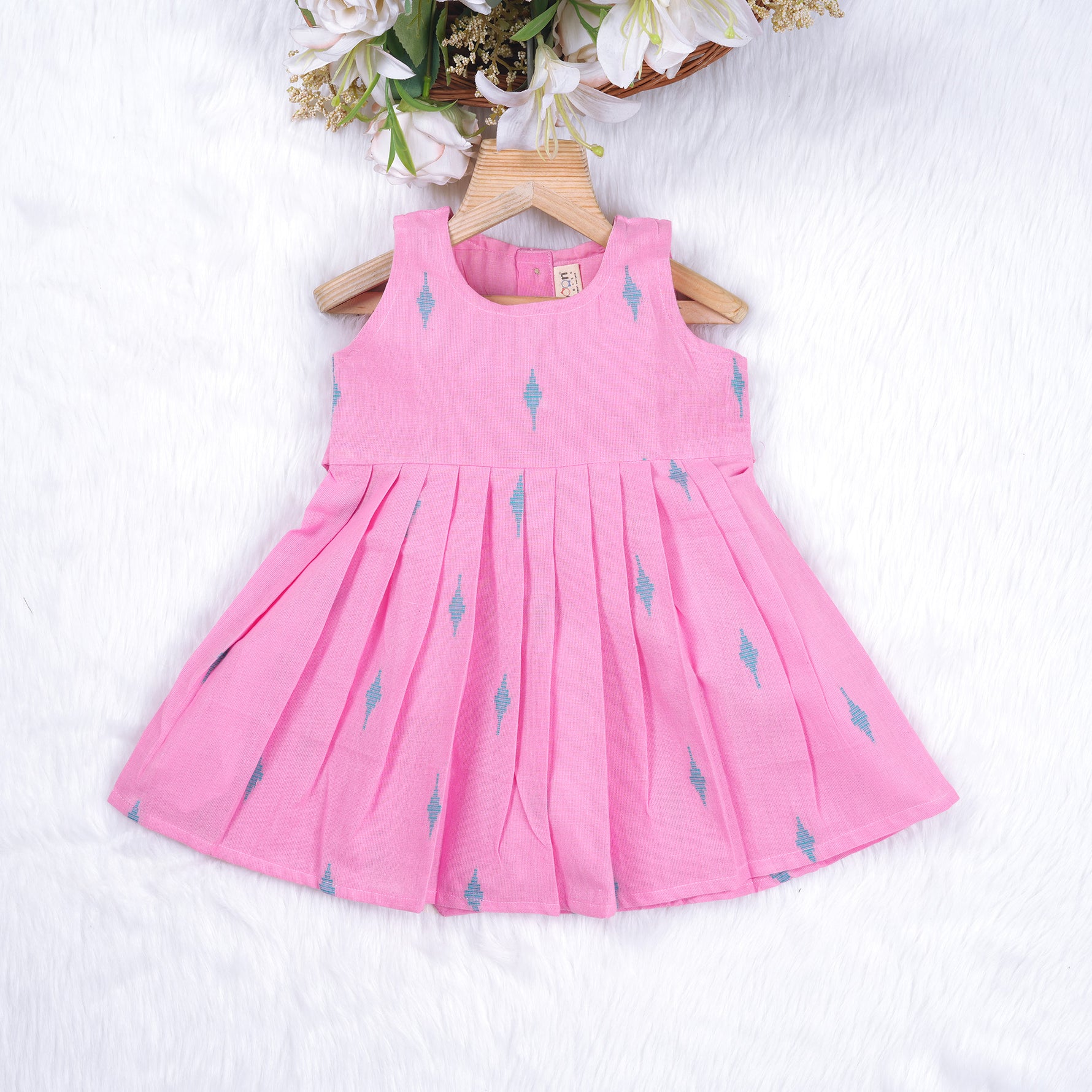 Pink sleeveless cotton frock with soft pleats and blue ikat motifs, displayed on a wooden hanger under a basket of white flowers – from Boon Babies girlswear collection.
