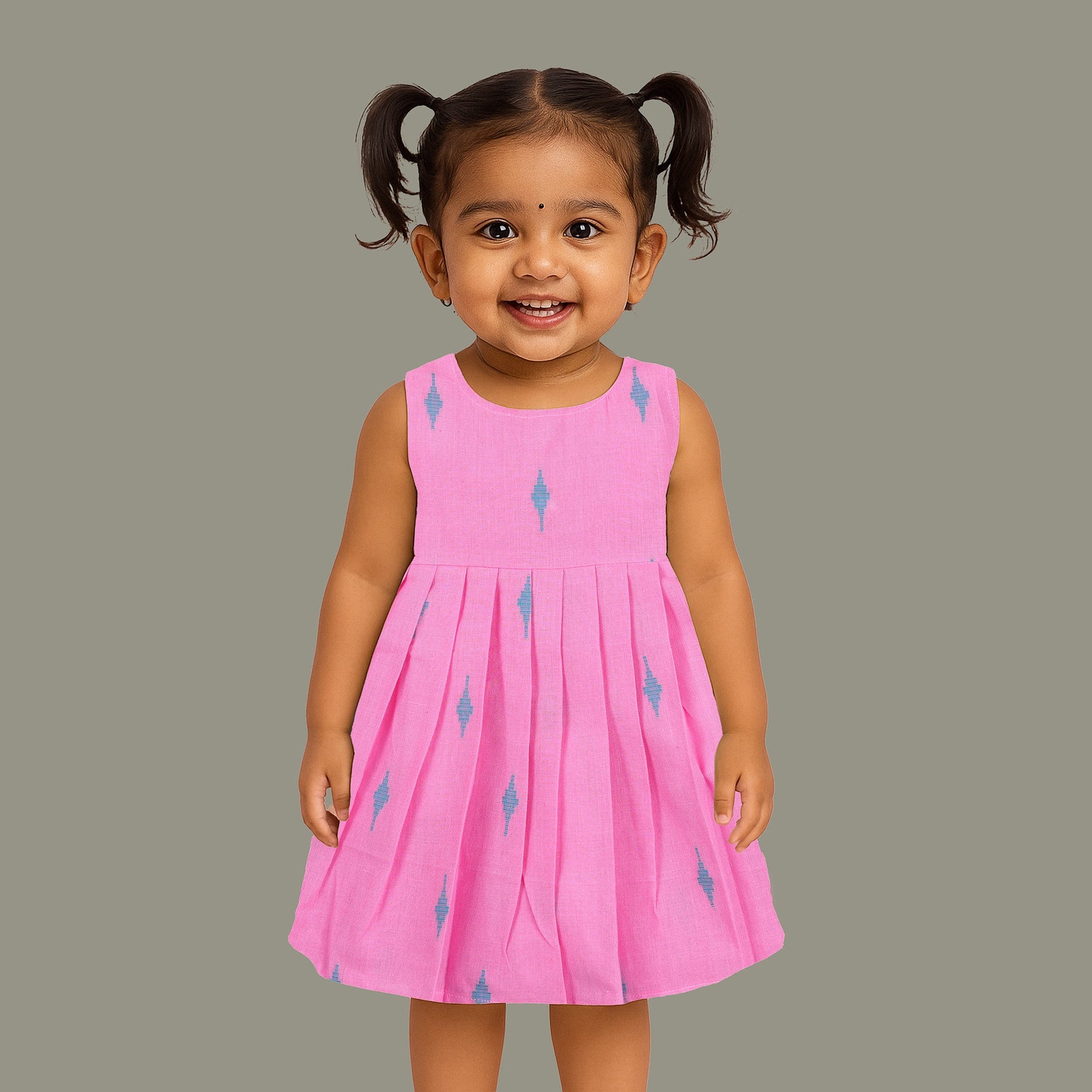 Pink sleeveless cotton frock with soft pleats and blue ikat motifs, displayed on a wooden hanger under a basket of white flowers – from Boon Babies girlswear collection.
