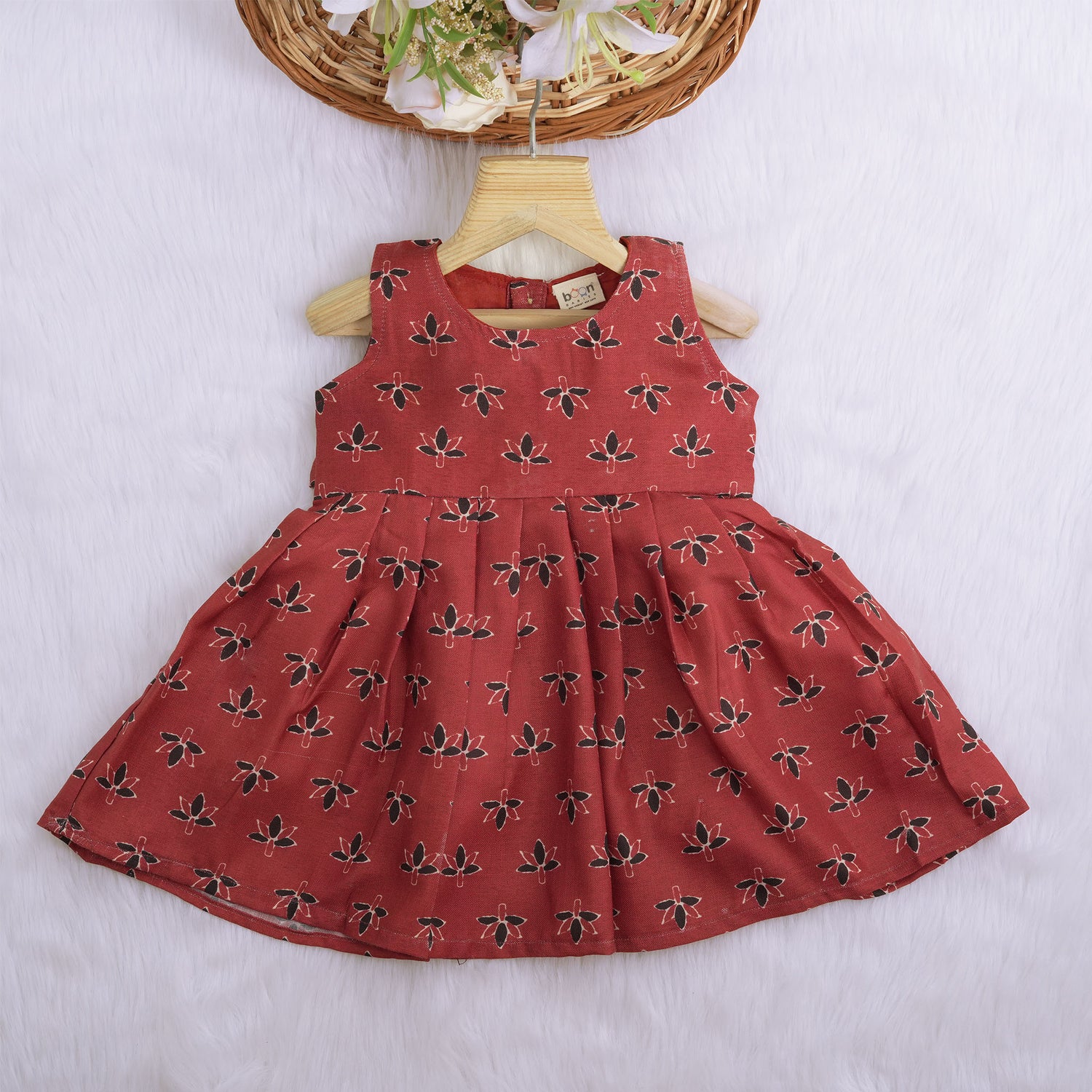Rust orange sleeveless slub cotton frock for baby girls with black floral prints and pleated skirt, displayed on a white fur background with floral basket hanger.