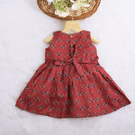 Rust orange sleeveless slub cotton frock for baby girls with black floral prints and pleated skirt, displayed on a white fur background with floral basket hanger.