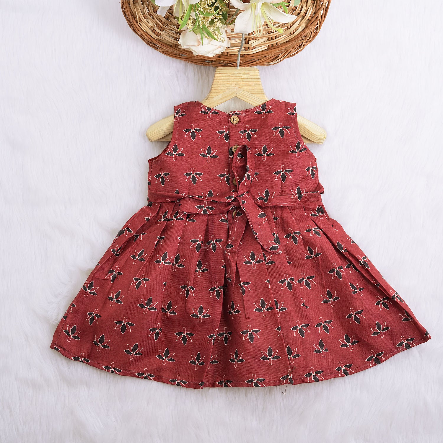 Rust orange sleeveless slub cotton frock for baby girls with black floral prints and pleated skirt, displayed on a white fur background with floral basket hanger.