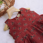 Rust orange sleeveless slub cotton frock for baby girls with black floral prints and pleated skirt, displayed on a white fur background with floral basket hanger.