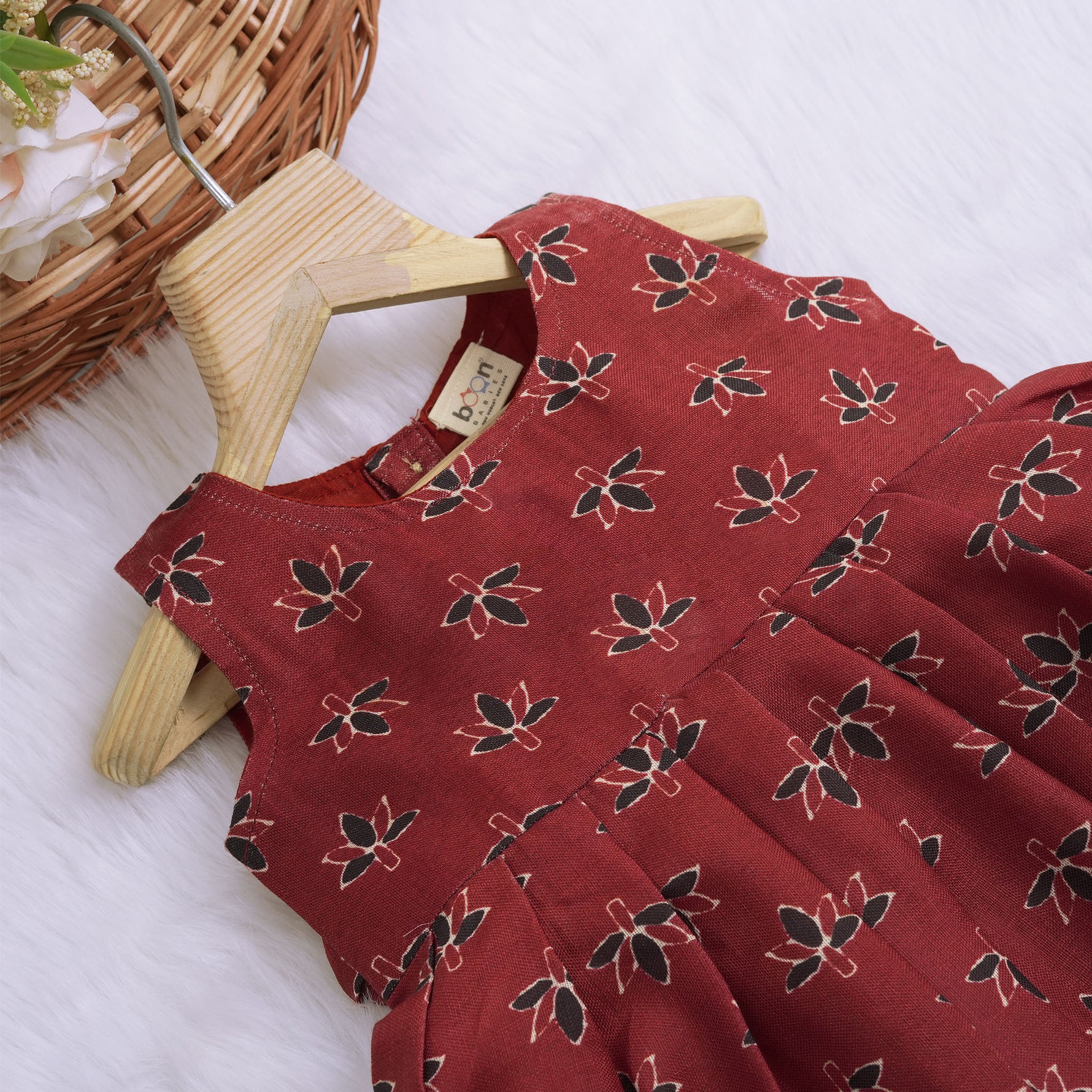 Rust orange sleeveless slub cotton frock for baby girls with black floral prints and pleated skirt, displayed on a white fur background with floral basket hanger.