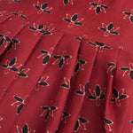 Rust orange sleeveless slub cotton frock for baby girls with black floral prints and pleated skirt, displayed on a white fur background with floral basket hanger.