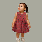 Rust orange sleeveless slub cotton frock for baby girls with black floral prints and pleated skirt, displayed on a white fur background with floral basket hanger.