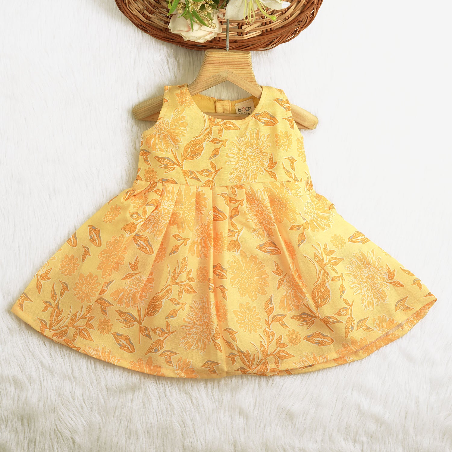 Sunshine yellow sleeveless slub cotton frock for baby girls with all-over orange floral print and pleated skirt, displayed flat on a white fur background with floral basket hanger.