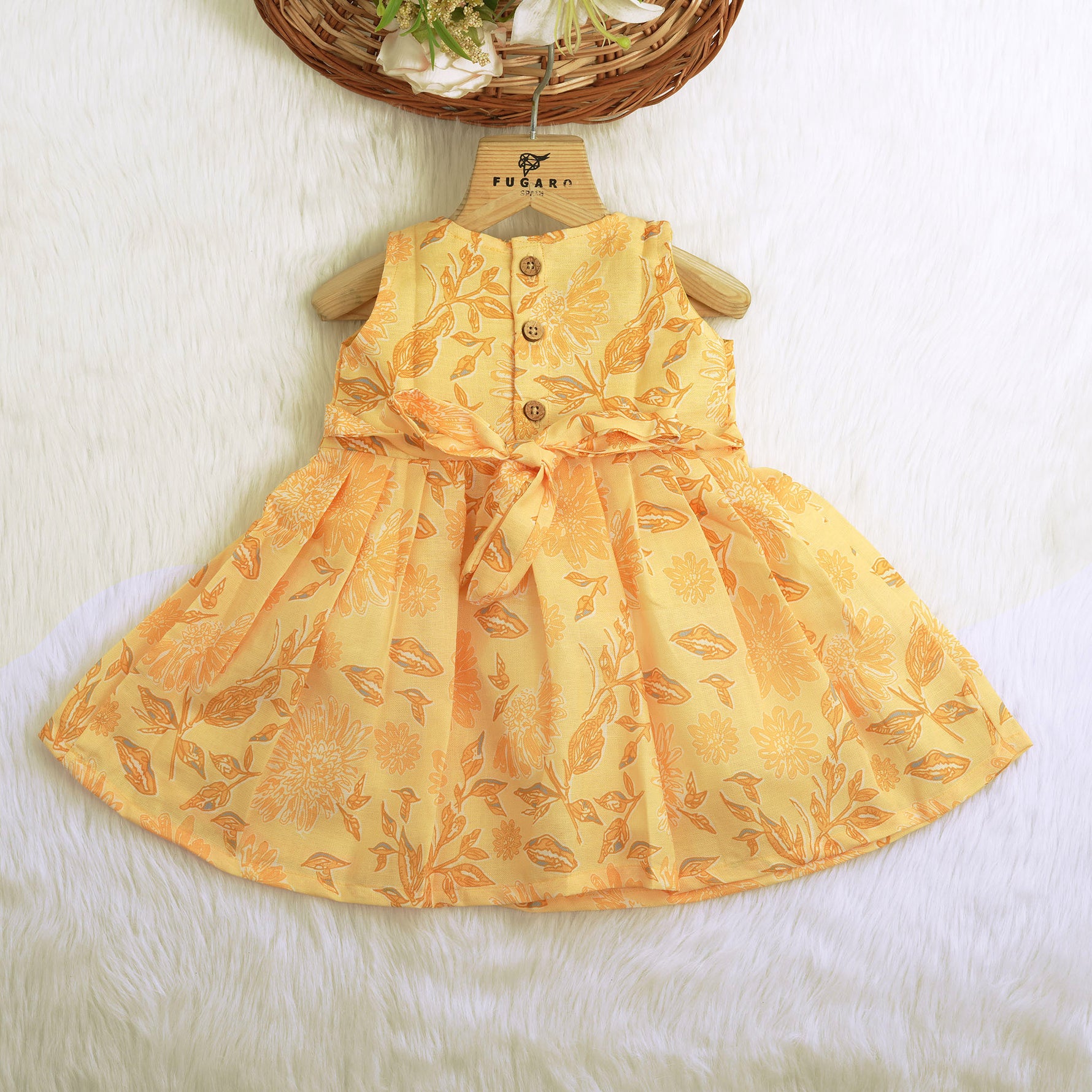 Sunshine yellow sleeveless slub cotton frock for baby girls with all-over orange floral print and pleated skirt, displayed flat on a white fur background with floral basket hanger.