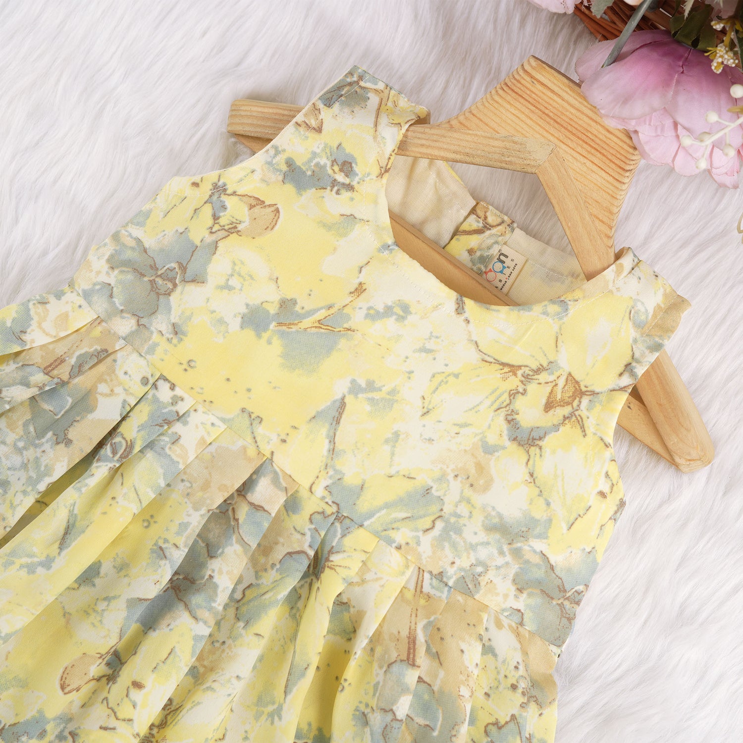 Yellow sleeveless floral chiffon cotton frock for baby girls, with pleated skirt and pastel tones, displayed on a wooden hanger over a white fur background and floral décor.