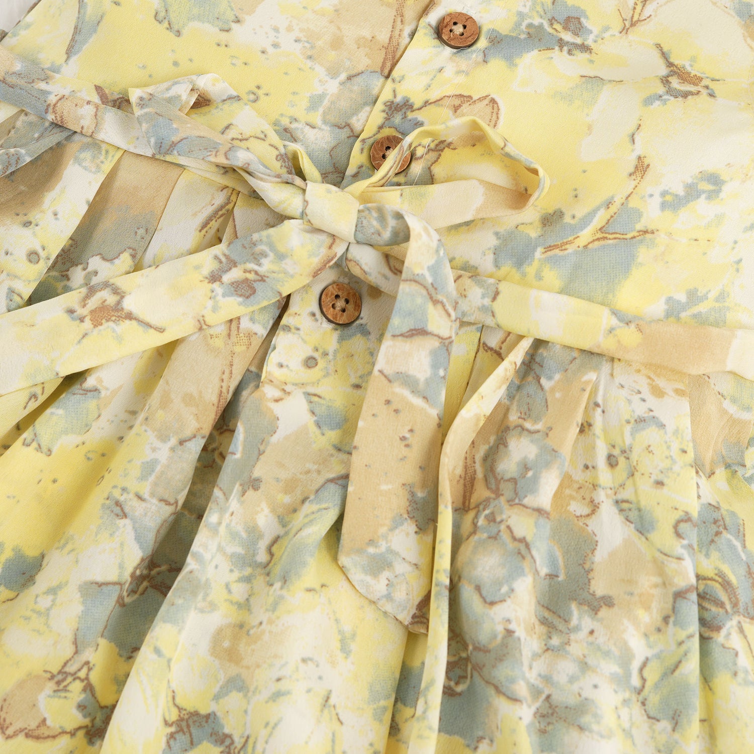 Yellow sleeveless floral chiffon cotton frock for baby girls, with pleated skirt and pastel tones, displayed on a wooden hanger over a white fur background and floral décor.