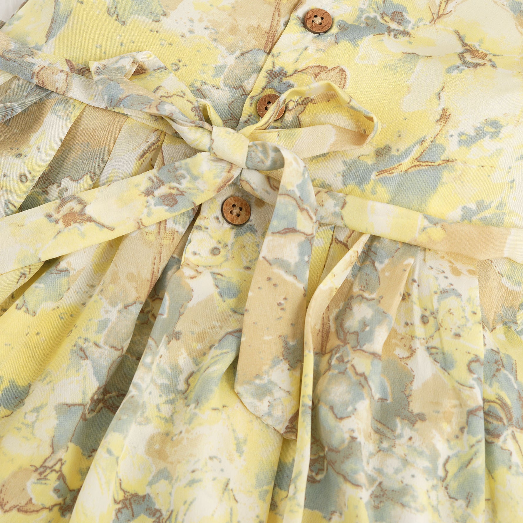 Yellow sleeveless floral chiffon cotton frock for baby girls, with pleated skirt and pastel tones, displayed on a wooden hanger over a white fur background and floral décor.
