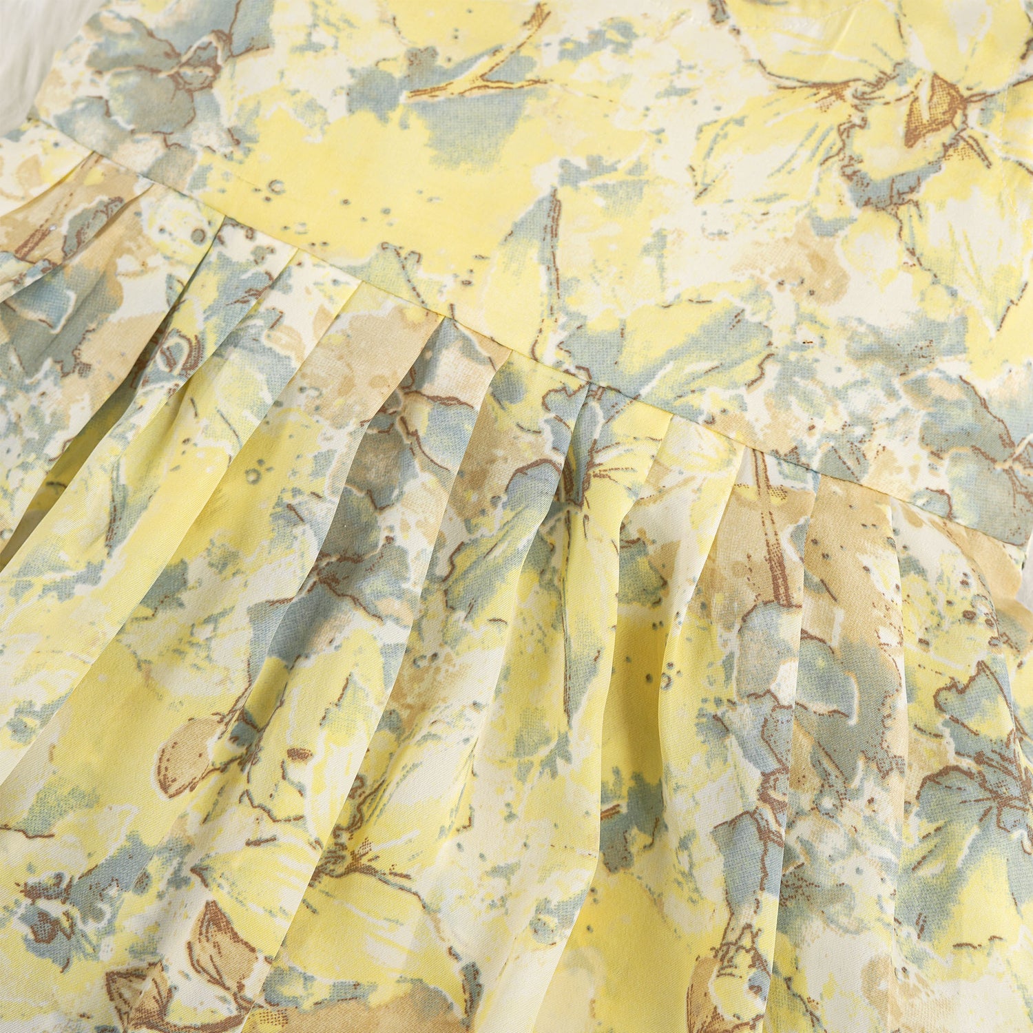 Yellow sleeveless floral chiffon cotton frock for baby girls, with pleated skirt and pastel tones, displayed on a wooden hanger over a white fur background and floral décor.