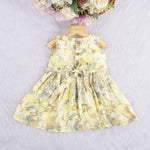 Yellow sleeveless floral chiffon cotton frock for baby girls, with pleated skirt and pastel tones, displayed on a wooden hanger over a white fur background and floral décor.