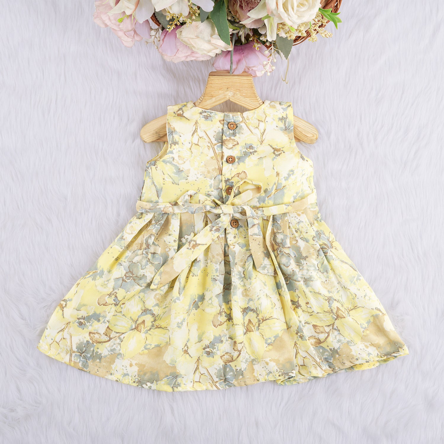 Yellow sleeveless floral chiffon cotton frock for baby girls, with pleated skirt and pastel tones, displayed on a wooden hanger over a white fur background and floral décor.