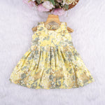Yellow sleeveless floral chiffon cotton frock for baby girls, with pleated skirt and pastel tones, displayed on a wooden hanger over a white fur background and floral décor.