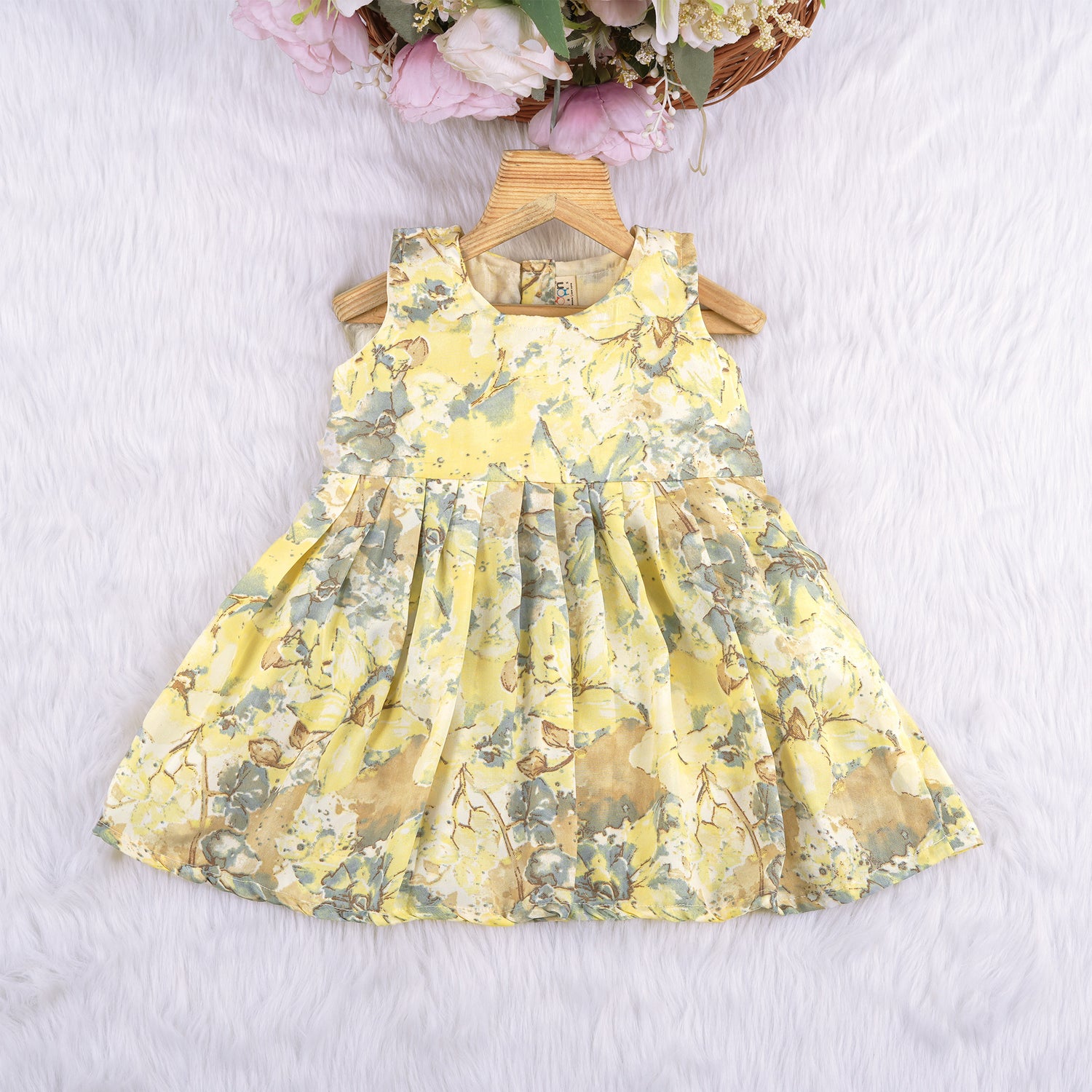 Yellow sleeveless floral chiffon cotton frock for baby girls, with pleated skirt and pastel tones, displayed on a wooden hanger over a white fur background and floral décor.