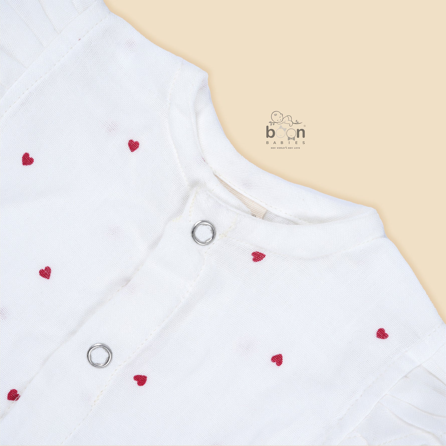 White muslin baby romper with tiny red heart prints, featuring frill sleeves, side bows, and front snap buttons – from Boon Babies, Coimbatore.