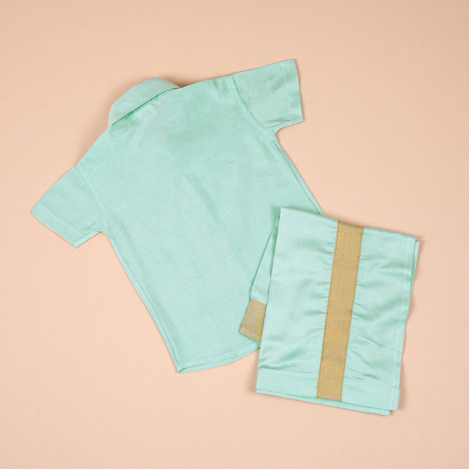 Teal blue silk dhoti shirt set for baby boys featuring a wooden-button shirt and gold-bordered dhoti, displayed flat on a beige background with floral props.
