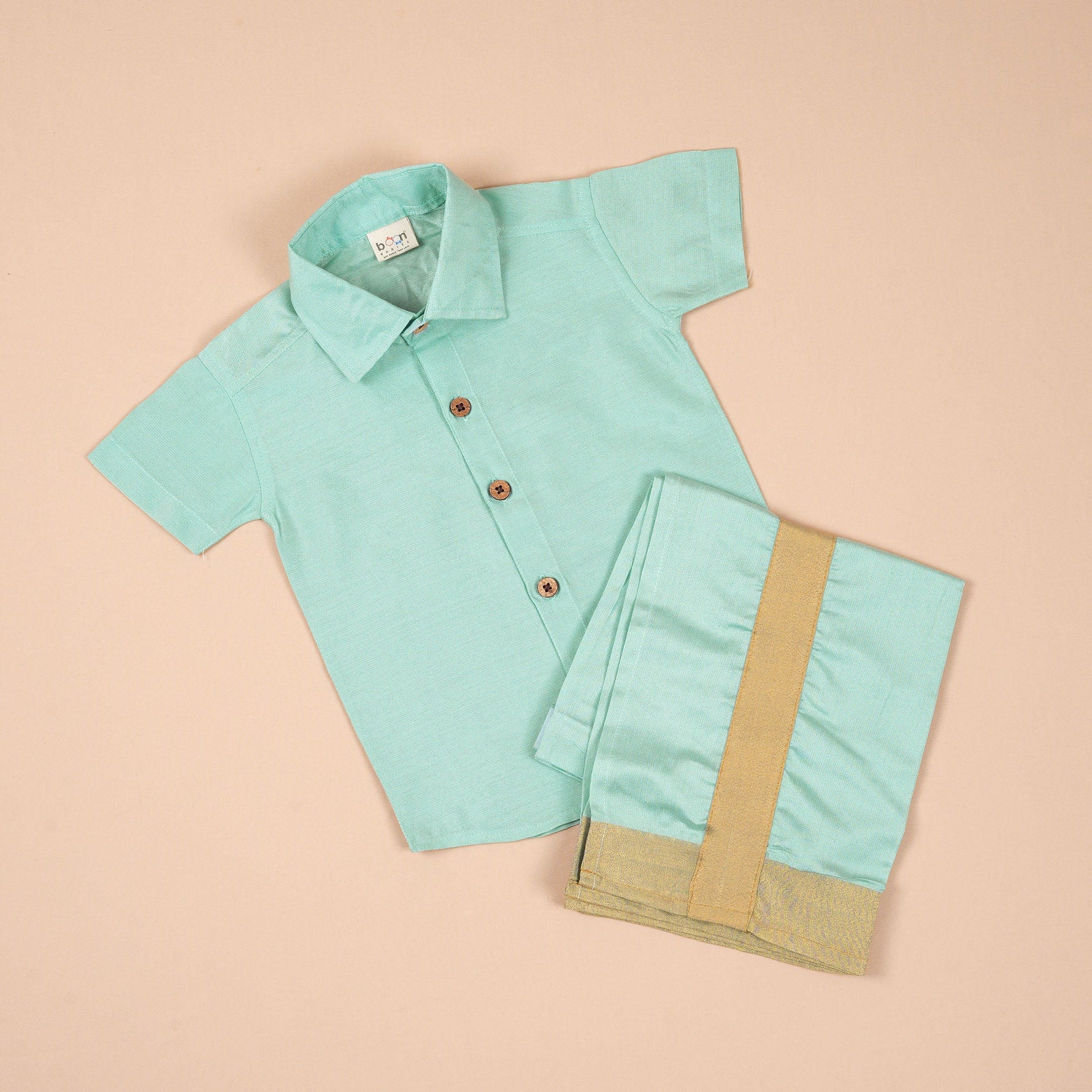 Teal blue silk dhoti shirt set for baby boys featuring a wooden-button shirt and gold-bordered dhoti, displayed flat on a beige background with floral props.